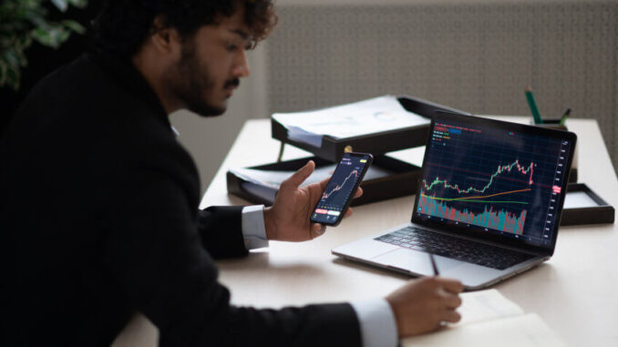 Smart focused Indian broker investor, stock trader using smartphone and laptop to analyze and invert in the financial market of cryptocurrencies, buy or sell crypto coins, planning strategy