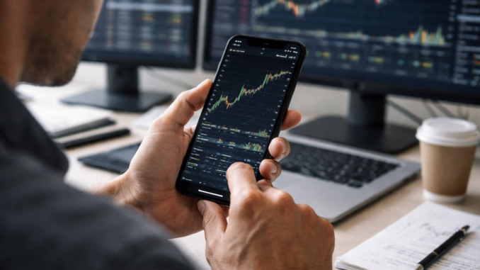 A trader analyzes a financial price chart on a smartphone while multiple market charts display on monitors in the background.